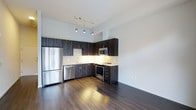 This single-floor space showcases a contemporary open-concept design, anchored by a well-appointed kitchen that serves as the focal point of the layout. The kitchen features dark, wood-grain cabinetry with a striking vertical stripe pattern, providing a bold visual contrast against the crisp white walls and bright subway tile backsplash. A full suite of stainless steel appliances — including a French door refrigerator, dishwasher, built-in microwave, and a freestanding range — offers both style and functionality. Under-cabinet lighting illuminates the countertops, enhancing the workspace and adding warmth to the overall aesthetic. Suspended above, a sleek track lighting system with modern pendant-style fixtures highlights the kitchen area while contributing to an elevated, polished look. The space flows seamlessly into an open living area, unified by rich, dark hardwood flooring that runs throughout, adding depth and sophistication. The high ceilings amplify the sense of openness, while the clean white walls keep the space feeling bright and airy. The overall ambiance is modern and refined, offering a functional yet stylish environment.