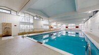 Located at 200 Rideau Terrace in Ottawa, Ontario, this expansive indoor aquatic facility spans a generous single floor and is purpose-built around a large rectangular lap pool. The pool itself features clearly marked depth indicators, ranging from a shallow entry area to a deep end of 8 feet, and is equipped with stainless steel entry ladders and poolside safety equipment including life rings and a rescue board.

The architecture is defined by soaring vaulted ceilings supported by exposed structural beams, lending the space a bright and open atmosphere. Floor-to-ceiling and clerestory windows line multiple walls, flooding the interior with natural light while offering views of the mature trees and green space surrounding the facility. The white ceramic tile walls and light-toned pool deck complement the airy aesthetic throughout.

Along one side of the pool, a continuous wooden bench runs the length of the space, providing seating for swimmers and observers alike. The pool deck offers generous circulation space on all sides. A glazed interior partition with double doors separates the pool hall from an adjacent entry or observation area, which features additional natural wood storage benches and safety equipment displays. Emergency exits with direct outdoor access are positioned at multiple points along the perimeter, ensuring both safety and connectivity to the surrounding grounds.