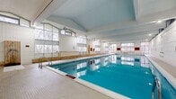 Located at 200 Rideau Terrace in Ottawa, Ontario, this expansive indoor aquatic facility spans a generous single floor and is purpose-built around a large rectangular lap pool. The pool itself features clearly marked depth indicators, ranging from a shallow entry area to a deep end of 8 feet, and is equipped with stainless steel entry ladders and poolside safety equipment including life rings and a rescue board.

The architecture is defined by soaring vaulted ceilings supported by exposed structural beams, lending the space a bright and open atmosphere. Floor-to-ceiling and clerestory windows line multiple walls, flooding the interior with natural light while offering views of the mature trees and green space surrounding the facility. The white ceramic tile walls and light-toned pool deck complement the airy aesthetic throughout.

Along one side of the pool, a continuous wooden bench runs the length of the space, providing seating for swimmers and observers alike. The pool deck offers generous circulation space on all sides. A glazed interior partition with double doors separates the pool hall from an adjacent entry or observation area, which features additional natural wood storage benches and safety equipment displays. Emergency exits with direct outdoor access are positioned at multiple points along the perimeter, ensuring both safety and connectivity to the surrounding grounds.