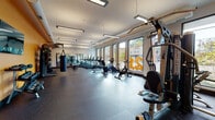 Located on Larchmere Boulevard in Cleveland's vibrant Larchmere neighborhood, this well-equipped fitness center offers a comprehensive workout environment on a single open floor. The space is thoughtfully designed with a clean, modern aesthetic, highlighted by large floor-to-ceiling windows along one wall that flood the interior with natural light and offer views of the surrounding streetscape and greenery.

The main workout floor features durable rubber flooring throughout and is divided into distinct functional zones. A dedicated cardio section houses a row of treadmills, ellipticals, stationary bikes, and a rowing machine, all positioned to take advantage of the expansive window views. A strength training area includes a multi-station cable machine and a variety of resistance equipment, while a free weights zone offers a full rack of dumbbells alongside a wall-mounted flat-screen television. A heavy punching bag adds a combat fitness option to the mix.

The facility is brightly lit by overhead linear pendant lighting that complements the natural daylight. A bold yellow accent wall adds energy and visual character to the space, contrasting with the neutral white walls and dark flooring. Full-length mirrors along one wall enhance the sense of openness. The overall ambiance is clean, functional, and motivating, making this fitness center a well-rounded training destination.