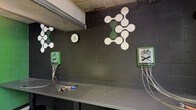 This single-floor space is designed as a dedicated bicycle repair and maintenance area. The room is defined by its dark-painted cinder block walls, which lend an industrial character to the environment. Two "Public Toolbox" stations are mounted on the wall, each clearly marked with bicycle repair iconography and equipped with tethered tools on cables, ensuring tools remain accessible and secure. A continuous work surface runs along the wall, providing ample space for bike maintenance tasks. A round wall clock is centrally mounted, offering a functional reference point within the space. Decorative molecular or atomic-style graphic elements in green and white are applied to the walls, adding a distinctive visual identity to the otherwise utilitarian setting. The ceiling features exposed structural elements, reinforcing the industrial aesthetic. The overall ambiance is practical and purposeful, clearly outfitted to serve cyclists in need of on-the-spot repairs, with an organized layout that keeps essential tools within easy reach.