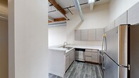 Located at 550 W Western Ave in Muskegon, Michigan, this one-bedroom, one-bathroom loft-style residence masterfully combines industrial character with contemporary updates. The open layout features soaring ceilings with exposed wooden beams, visible ductwork, and wood-look plank flooring throughout the main living and kitchen areas, creating an authentic urban loft aesthetic.

The modern kitchen is a standout feature, outfitted with sleek flat-front cabinetry in a warm gray tone, white quartz countertops, a peninsula with an undermount sink, and a full suite of stainless steel appliances including a refrigerator, electric range with microwave, and dishwasher. The high ceilings in the kitchen amplify the sense of openness.

The living area showcases original exposed brick walls and large, wood-framed windows that flood the space with natural light and offer views of the surrounding cityscape. Plush carpeting anchors the room with warmth. The bedroom continues the industrial-chic design with soaring ceilings, exposed beams, and soft carpeting, while two closets provide generous storage, one of which features original brick detailing. The full bathroom includes a combination tub and shower. This space is a compelling blend of historic character and modern convenience.