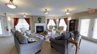 This single-floor space exudes a warm, inviting ambiance anchored by a classic white-mantled gas fireplace that serves as the focal point of the main living area. The room is generously proportioned, accommodating multiple seating arrangements including plush gray sofas, striped wingback chairs, and a central wooden coffee table — all thoughtfully arranged to encourage conversation and relaxation. A patterned area rug adds a touch of color and definition to the carpeted floor. Natural light pours in through multiple windows dressed in rich, deep red curtains, creating a striking contrast against the neutral beige and sage green walls. Flush-mount ceiling light fixtures with decorative pendants are evenly distributed throughout, providing ample overhead lighting to complement the natural brightness. Wall sconces flank the fireplace, adding a refined finishing touch. A dark wood bookcase in the corner provides both storage and display space, lending a homey, lived-in character to the room. The overall aesthetic blends traditional design elements with comfortable, residential furnishings, resulting in a space that feels both functional and elegantly understated.