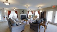 This single-floor space exudes a warm, inviting ambiance anchored by a classic white-mantled gas fireplace that serves as the focal point of the main living area. The room is generously proportioned, accommodating multiple seating arrangements including plush gray sofas, striped wingback chairs, and a central wooden coffee table — all thoughtfully arranged to encourage conversation and relaxation. A patterned area rug adds a touch of color and definition to the carpeted floor. Natural light pours in through multiple windows dressed in rich, deep red curtains, creating a striking contrast against the neutral beige and sage green walls. Flush-mount ceiling light fixtures with decorative pendants are evenly distributed throughout, providing ample overhead lighting to complement the natural brightness. Wall sconces flank the fireplace, adding a refined finishing touch. A dark wood bookcase in the corner provides both storage and display space, lending a homey, lived-in character to the room. The overall aesthetic blends traditional design elements with comfortable, residential furnishings, resulting in a space that feels both functional and elegantly understated.