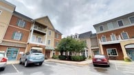 This single-floor unit is situated within a well-established mixed-use development characterized by traditional architectural styling. The exterior of the building showcases a combination of brick and painted siding, multi-paned windows with shutters, covered balconies, and pitched rooflines with dormers — all contributing to a classic, neighborhood-friendly aesthetic. Mature landscaping, including a prominent shade tree, anchors the central courtyard and enhances the building's welcoming curb appeal.

Inside, the unit maintains a clean, neutral palette throughout, with white walls and light-toned carpeting that create a bright and airy atmosphere. The bedroom is a generously proportioned space, illuminated by a ceiling fan with a light fixture, and features two doorways — one leading to an en-suite bathroom and the other opening to the broader unit. A dedicated walk-in closet provides substantial storage, fitted with a full-width hanging rod and carpeted flooring for comfort underfoot.

A hallway area benefits from multiple windows dressed with white horizontal blinds, allowing natural light to filter through while maintaining privacy. The overall layout is straightforward and functional, well-suited for comfortable single-level living within a vibrant, pedestrian-friendly community setting.
