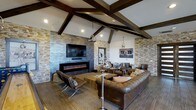 This single-floor space blends rustic character with contemporary design in a seamlessly curated interior. The main living area commands attention with its vaulted ceiling adorned by bold, dark-stained exposed wood beams that draw the eye upward and lend a dramatic architectural quality to the room. Stacked stone accent walls in warm, earthy tones frame the space, adding rich texture and a sense of natural craftsmanship throughout.

A sleek, linear electric fireplace serves as a focal point, set beneath a mounted flat-screen television against a sophisticated dark tile surround — a striking contrast to the lighter stone walls on either side. Pendant lighting in a polished industrial style hangs from the beamed ceiling, casting warm, ambient light across the room.

The furnishings are equally thoughtful, featuring generously sized leather sofas and chairs in warm browns, complemented by a solid wood coffee table that reinforces the space's organic, lodge-inspired aesthetic. Wide-plank wood-look flooring grounds the entire room with continuity and warmth.

Overall, the space exudes a welcoming, upscale ambiance — a refined retreat that balances natural materials with modern comforts, making it well-suited for both relaxed gatherings and impressive entertaining.