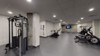 This fitness center offers a comprehensive exercise environment spread across a single open floor. The space features a drop tile ceiling with recessed lighting throughout, creating consistent, even illumination across all workout zones. Durable gray rubber flooring covers the entire area, providing both safety and functionality for various exercise activities.

The cardio section is well-appointed with multiple air bikes, elliptical trainers, and a treadmill, positioned to allow comfortable spacing between machines. A wall-mounted flat-screen television is visible from this zone, providing entertainment during workouts. The strength training area includes a multi-station cable machine, a dumbbell rack with a full range of weights, and adjustable workout benches, catering to a variety of resistance training needs.

A dedicated functional training zone features exercise mats laid out for floor-based workouts, alongside a curated selection of kettlebells, medicine balls, and plyo boxes neatly organized on a storage rack. A workout reference chart is mounted on the wall nearby for guidance.

Exposed concrete columns add an industrial character to the space, complementing the clean white walls. The layout is open and uncluttered, allowing for smooth circulation and simultaneous use of all equipment zones.