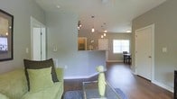 This single-floor space features an open-concept layout that seamlessly connects the living, kitchen, and dining areas. The living room is anchored by rich, dark hardwood flooring and neutral gray walls, creating a clean, modern backdrop. A glass-top coffee table and a light green sofa add a fresh, contemporary feel to the space.

A half-wall kitchen bar with a granite-style countertop divides the living area from the kitchen, offering both visual separation and functional workspace. Pendant lights hang in a cluster above the bar, providing focused task lighting and a stylish focal point. The kitchen features a standard faucet setup visible above the counter.

Beyond the kitchen bar, the space opens further into a dining area, illuminated by natural light streaming through a prominent window. The warm glow from the window brightens the entire open-plan area, enhancing the sense of spaciousness. Track lighting near the entryway adds additional illumination, while a flush-mount ceiling fixture serves the dining zone. White trim and baseboards throughout provide crisp contrast against the neutral wall tones, contributing to the space's polished, cohesive aesthetic.