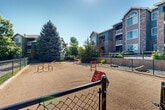 Located at 7927 W Mansfield Pkwy in Lakewood, Colorado, this outdoor dog park is a dedicated amenity within a well-established residential community. The park features a spacious, level sandy surface enclosed by a chain-link fence with a gated entry, providing a safe and contained area for pets to roam freely. Several colorful agility obstacles are distributed throughout the space, including hurdle jumps, hoop tunnels, and weave poles, offering engaging activities for dogs of various sizes and skill levels. The perimeter is bordered by decorative rock landscaping, ornamental boulders, and mature trees that provide shade and a natural aesthetic. Surrounding the park on multiple sides are multi-story residential buildings constructed with brick and siding exteriors, featuring private balconies and arched windows that overlook the greenspace. Lush lawns, shrubs, and carefully maintained plantings frame the community backdrop. The open layout and clear Colorado skies contribute to a bright, welcoming atmosphere. Benches nearby offer a comfortable spot for pet owners to relax while their dogs enjoy the agility course. The thoughtful design of this dog park reflects a community-oriented approach to outdoor amenity spaces.