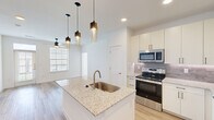 Located in Manor, TX, this thoughtfully designed 1-bedroom, 1-bathroom residence offers a bright, modern aesthetic across its well-appointed layout. The open-concept living and kitchen area is anchored by light wood-tone flooring, crisp white walls, and abundant natural light streaming through large windows. The kitchen is a standout feature, showcasing a granite-topped center island with an undermount sink, stylish pendant lighting, white shaker cabinetry, a subway tile backsplash, and a full suite of stainless steel appliances including a refrigerator with ice maker, range, microwave, and dishwasher. Built-in shelving near the entry adds functional storage and visual interest. The living area extends naturally from the kitchen, enhanced by a ceiling fan and access to a private balcony overlooking a serene pond and landscaped community grounds. The bedroom offers plush carpet flooring, a ceiling fan, and a window with outdoor views. It connects to a spacious walk-in closet outfitted with multi-tier shelving and hanging rods. The bathroom features granite countertops, white cabinetry with modern hardware, a framed mirror, and a tiled walk-in shower.