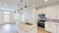 Located in Manor, TX, this thoughtfully designed 1-bedroom, 1-bathroom residence offers a bright, modern aesthetic across its well-appointed layout. The open-concept living and kitchen area is anchored by light wood-tone flooring, crisp white walls, and abundant natural light streaming through large windows. The kitchen is a standout feature, showcasing a granite-topped center island with an undermount sink, stylish pendant lighting, white shaker cabinetry, a subway tile backsplash, and a full suite of stainless steel appliances including a refrigerator with ice maker, range, microwave, and dishwasher. Built-in shelving near the entry adds functional storage and visual interest. The living area extends naturally from the kitchen, enhanced by a ceiling fan and access to a private balcony overlooking a serene pond and landscaped community grounds. The bedroom offers plush carpet flooring, a ceiling fan, and a window with outdoor views. It connects to a spacious walk-in closet outfitted with multi-tier shelving and hanging rods. The bathroom features granite countertops, white cabinetry with modern hardware, a framed mirror, and a tiled walk-in shower.