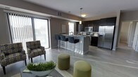 This single-floor residence showcases a thoughtfully designed open-concept layout that seamlessly connects the living and kitchen areas, creating a spacious and cohesive living environment. The kitchen is a standout feature, boasting rich dark cabinetry, granite countertops, and stainless steel appliances including a French door refrigerator, range, and microwave. A large central island with bar seating provides both additional prep space and a casual dining option, illuminated by pendant lighting overhead. The light-toned hardwood-style flooring runs throughout, lending a clean and contemporary aesthetic to the space. The living area is warm and welcoming, featuring a patterned accent chair and olive green upholstered ottomans that add a pop of color against the neutral gray walls. A large sliding glass door floods the space with natural light and offers views to an outdoor balcony or patio area, further enhancing the open and airy feel. Decorative greenery adds a refreshing, organic touch to the overall design. The combination of modern finishes, neutral tones, and functional layout makes this residence a refined and comfortable place to call home.