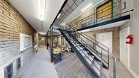 Located at 15111 Greenfield Rd in Detroit, Michigan, this two-story building offers a distinctive architectural character defined by exposed light-colored brick walls, bold black steel structural elements, and an enclosed interior courtyard that serves as the heart of the space. The courtyard atrium is accessible from both floors and is furnished with picnic tables and decorative planters, creating a communal gathering area beneath a soaring open ceiling structure that floods the space with light.

The ground floor hallway connects unit entrances along the brick facade, with large windows providing visibility into the courtyard. The upper floor mirrors this layout, with a walkway corridor featuring sturdy black steel railings that overlook the atrium below. A prominent steel staircase with open risers and geometric railing details connects the two levels, functioning as both a practical and visual centerpiece of the building.

The upper level hallway features a suspended grid ceiling with recessed lighting panels, white-painted walls complementing the raw brick accent wall, and wide windows that bring in natural light. Throughout both floors, the interplay of industrial materials — steel, brick, and concrete — creates a utilitarian yet characterful ambiance. The building's layout across two comparably sized floors provides a balanced and functional flow throughout.