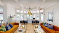 Located at 5010 Central Sarasota Pkwy in Sarasota, Florida, this single-floor community clubhouse offers a thoughtfully designed and generously proportioned amenity space. The reception area serves as a stunning focal point, featuring a marble-accented front desk beneath a dramatic cluster of amber pendant lights. Floor-to-ceiling windows wrap around the curved reception room, flooding the space with natural light and offering direct views of the outdoor pool area.

The open interior flows into a vibrant lounge featuring coffered ceilings, arched doorways with decorative columns, and light hardwood flooring throughout. Bold orange velvet sofas and gold-framed glass coffee tables add a touch of eclectic sophistication. Adjacent to the lounge, a spacious game room is anchored by two pool tables with gold metal frames set atop deep blue area rugs. Window seats with floral-patterned Roman shades and botanical wallpaper lend a refined, resort-like atmosphere.

The dining area is a design standout, featuring a striking blue geometric accent wall with sculptural paneling, a round white dining table, woven leather chairs, and a warm wood slat room divider. A separate bar-height counter with wicker seating offers additional gathering space. Together, these areas create a welcoming, design-forward clubhouse ideal for socializing and recreation.