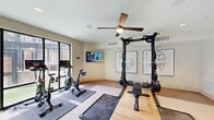 This fitness center offers a comprehensive, well-appointed exercise environment across a single generously sized floor. The space is thoughtfully designed with a neutral color palette of warm whites and beige tones, complemented by light carpet and hardwood flooring sections that define distinct workout zones.

The cardio area is positioned along a dramatic floor-to-ceiling window wall, allowing abundant natural light and views of the surrounding exterior. It is equipped with treadmills, a stair climber, and additional cardio machines, all facing outward for an engaging workout experience. Ceiling fans with stylish gold and brushed finishes are installed throughout, ensuring air circulation across the entire space.

The strength training section features a Matrix power rack, adjustable benches, a full dumbbell rack with a wide range of weights, and a cable machine — all mirrored by elegant arched full-wall mirrors that visually expand the room. A dedicated spin bike and mat area with a wall-mounted TV provides a defined studio-style zone, decorated with equestrian line-art prints for a refined aesthetic touch.

A built-in wet bar or refreshment station with cabinetry and a sink is tucked into an alcove near the entrance, adding a practical amenity. Recessed lighting and ceiling speakers complete the polished, functional atmosphere of this fully outfitted fitness facility.