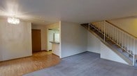This two-story residence offers a practical and straightforward layout designed for comfortable everyday living. The main level features an open living area finished with wall-to-wall carpeting, which flows seamlessly into a hardwood-floored entryway near the front door. The kitchen is positioned toward the rear of the main floor and is equipped with white cabinetry, a white refrigerator, and a raised breakfast bar that provides a natural divide between the kitchen and the living space. The neutral, light-toned walls throughout the main level create a clean and airy atmosphere. A staircase with a metal railing leads from the main level to the upper floor, adding a sense of vertical openness to the space. The overall design is functional and uncluttered, making it well-suited for a variety of living arrangements. The combination of hardwood flooring in the entryway and carpeting in the living area adds a subtle variation in texture and material that defines each zone of the main level. The upper floor provides additional living space, accessible via the centrally located staircase.