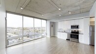 This contemporary 1-bedroom, 1-bathroom residence showcases an open-concept floor plan that seamlessly blends living and kitchen areas into a cohesive, functional space. The industrial-inspired design is defined by exposed concrete waffle ceilings, which lend an urban edge and architectural character throughout. Light-toned hardwood flooring runs continuously underfoot, complementing the bright, airy atmosphere.

Expansive floor-to-ceiling windows along the corner of the living area flood the space with natural light and frame sweeping panoramic views of the surrounding cityscape, creating a dynamic connection between interior and exterior. The kitchen is outfitted with crisp white cabinetry, a classic subway tile backsplash, dark stone countertops, and stainless steel appliances — including a range and built-in microwave — offering a clean, modern aesthetic. A deep sink with an urban view backdrop adds a distinctive touch.

Recessed lighting is strategically placed throughout, ensuring well-distributed illumination to complement the abundant natural light. The overall ambiance is sleek and sophisticated, balancing industrial elements with modern finishes to create a refined yet comfortable urban living environment.