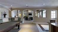 This single-story space showcases a thoughtfully designed interior with a warm, neutral color palette of taupe and white throughout. The layout is defined by distinctive arched doorways that elegantly connect multiple areas, creating a sense of openness while maintaining defined spaces. Recessed lighting and decorative pendant fixtures provide layered illumination across the rooms.

The foreground features a game room area with a full-size billiard table positioned on patterned tile flooring, which transitions into carpeted areas beyond. Through the arched openings, a formal lounge or sitting room is visible, complete with a fireplace serving as a focal point, a wall-mounted television, and traditional upholstered seating arranged around accent tables. Table lamps add a warm ambiance to this area.

Large windows in the lounge area allow natural light to flow into the space, with curtain panels framing the views. Crown molding and white trim detailing throughout add a refined architectural touch. The overall ambiance is polished and welcoming, combining recreational functionality with comfortable, lounge-style living areas suited for social gatherings.