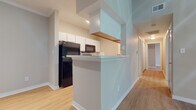 This single-floor residence is thoughtfully laid out to maximize both function and comfort. The kitchen is designed in a galley style, featuring white cabinetry with open shelving, light granite-style countertops, and black appliances including a full-size refrigerator, microwave, and dishwasher. A peninsula with a breakfast bar counter extends into the living area, creating a natural divide between the kitchen and the main living space while maintaining an open, connected feel. Light wood-tone flooring runs consistently throughout the entire unit, lending warmth and visual continuity to every room.

The living area is generously proportioned, benefiting from soaring vaulted ceilings and a ceiling fan with integrated lighting. Large windows and a glass-panel exterior door flood the space with natural light and provide views of surrounding greenery. A hallway connects the living area to the private rooms, each of which features the same light hardwood flooring, neutral-toned walls, and ceiling fans. Bedrooms are well-sized, with windows offering additional natural light and outdoor views.

The bathroom is clean and functional, featuring a white vanity with under-sink storage, a large mirror with overhead lighting, and a shower-tub combination. The overall ambiance of the space is bright, airy, and move-in ready.