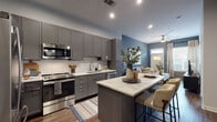 This single-floor residence showcases a thoughtfully designed open-concept layout with contemporary finishes throughout. The kitchen is a standout feature, equipped with full-height gray shaker cabinets, a white subway tile backsplash, stainless steel appliances including a range, microwave, dishwasher, and refrigerator, and a central island with a white quartz countertop that provides additional seating and prep space. Wood-look flooring runs throughout the main living areas, complementing the warm yet modern aesthetic. The kitchen flows seamlessly into the living room, which features a bold slate-blue accent wall, ceiling fan, and a bright entry door with windows that invite natural light. The primary bedroom continues the blue accent wall theme, offering a walk-in closet and rich hardwood-style flooring. A second bedroom features a large window with a street-facing view, bathing the room in natural light. The bathroom is finished with large-format white tile flooring, a dark-toned vanity with a white countertop, a wide mirror, and a fully tiled walk-in glass shower. Near the entry, a practical wall-mounted hook area provides a functional drop zone. The space presents a cohesive, modern design throughout.