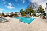 Located at University Station Circle in Charlotte, NC, this community amenity space centers around a generously sized outdoor swimming pool framed by an expansive paver deck. The pool area is well-appointed with multiple lounge chairs and seating arrangements, offering residents ample space to relax and socialize under the open sky. The surrounding landscape is thoughtfully designed, featuring mature magnolia trees, tall columnar evergreens, manicured hedges, ornamental grasses, and decorative stone planters filled with seasonal flowers, creating a lush and inviting atmosphere. A low iron fence defines the perimeter of the pool enclosure, maintaining a sense of privacy while preserving views of the broader community.

Adjacent to the pool, a single-story clubhouse building features a crisp white exterior with gray brick detailing, a pitched shingle roof, and large floor-to-ceiling windows that allow clear views of the pool from inside. The building's facade is accented by wall-mounted light fixtures and wide glass doors, blending functionality with a clean architectural aesthetic. The paver patio connecting the clubhouse to the pool deck provides additional outdoor seating areas with patio furniture, making the space ideal for casual gatherings. Overall, this amenity area offers a well-maintained, resort-like outdoor environment.