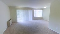 This single-floor space features a generously sized main living area finished with wall-to-wall carpeting in a neutral tone, creating a soft and comfortable foundation throughout the room. The space is characterized by an abundance of natural light, drawn in through a large set of floor-to-ceiling vertical blinds that appear to cover a sliding glass door or oversized window, alongside an additional wide window positioned to the right. Together, these openings provide views of surrounding greenery and flood the interior with bright, diffused daylight. The walls are painted in a soft, light neutral tone that complements the carpet and enhances the overall sense of openness. Structural beams are subtly visible along the ceiling, adding a modest architectural detail to the otherwise clean and understated design. A ceiling-mounted light fixture provides additional illumination. White baseboard trim lines the perimeter of the room, offering a crisp contrast to the carpeted floor. The space presents itself as move-in ready, with a simple, functional layout well-suited for a variety of furniture arrangements.