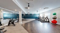 Located at 5391 W Fayetteville Rd in College Park, GA, this fitness center offers a thoughtfully designed exercise space spread across a single, generously sized floor. The room features a clean, modern aesthetic defined by a striking teal accent wall that contrasts beautifully with the neutral gray and white tones throughout. Durable wood-look flooring spans the entire space, transitioning subtly between lighter and darker tones to visually define different workout zones.

The cardio area is equipped with a row of treadmills positioned along the windowed wall, allowing natural light to filter in through slatted blinds. A dedicated cycling section lines the mirrored wall, where multiple stationary bikes are arranged for group or individual use. The floor-to-ceiling mirrors along one wall visually expand the space and are essential for form-checking during workouts.

A free weights section features a full dumbbell rack alongside an adjustable workout bench, while stability balls in multiple sizes add versatility for core and functional training. Multiple ceiling fans are mounted throughout, promoting consistent airflow across the open floor plan. Recessed lighting provides bright, even illumination throughout the space. The fitness center is accessible via a dark-framed entry door, with a water station conveniently located near the entrance.