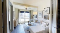 This single-floor residence exudes a clean, contemporary aesthetic designed for comfortable modern living. The living area serves as the heart of the space, bathed in abundant natural light streaming through large floor-to-ceiling windows dressed with elegant sheer and navy curtains. These expansive windows frame striking urban views, connecting the interior seamlessly to the cityscape beyond.

The living room is thoughtfully arranged with a plush, neutral-toned sectional sofa accented by patterned throw pillows, centered around a sleek glass and metallic coffee table. Wide-plank hardwood flooring runs throughout, lending warmth and continuity to the space. A ceiling fan with a brushed gold finish adds both function and a subtle decorative touch overhead.

Ambient and task lighting is well-considered, with a sculptural arc floor lamp providing a soft, inviting glow to complement the natural light. Curated wall art and framed prints add a sophisticated, gallery-like quality to the walls. A wooden sideboard near the entry area provides stylish storage, topped with tasteful decorative accents.

Overall, the space strikes a balance between casual comfort and refined style, making it an inviting and functional environment well-suited for both relaxing and entertaining.
