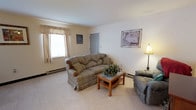 This single-floor space offers a straightforward, functional layout designed for comfortable everyday living. The living area serves as the heart of the home, featuring plush, plaid-upholstered seating including a generously sized sofa and a cushioned recliner chair, arranged around a wooden coffee table with a lower shelf for storage. The room is finished with neutral wall-to-wall carpeting that adds warmth underfoot, while the cream-colored walls provide a clean, bright backdrop throughout. A window dressed with a valance curtain allows natural light to filter into the space, enhancing its inviting atmosphere. Baseboard heating runs along the walls, ensuring year-round comfort. Decorative touches, including framed wall art and a classic table lamp, contribute to the lived-in, homey character of the space. A interior door provides access to additional areas of the home. Overall, this space presents a relaxed and practical environment, well-suited for everyday use with its approachable design and comfortable furnishings.