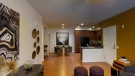 This single-story residence showcases an open-concept floor plan that seamlessly integrates the kitchen, dining, and living areas into one cohesive and functional space. Warm hardwood flooring flows throughout, lending a sense of continuity and warmth across the entire layout.

The kitchen is well-appointed with rich, dark wood cabinetry that extends to the upper walls, providing ample storage. A peninsula with a dark countertop and an undermount sink serves as both a functional workspace and a natural divider between the kitchen and the adjoining areas. Black appliances, including a refrigerator and microwave, complement the cabinetry tones.

The dining area is positioned centrally within the open plan and features a round table with upholstered chairs, anchored beneath a large framed landscape artwork that adds a sophisticated focal point to the space. Decorative circular wall accents on the adjacent wall introduce a textural, artistic element.

Recessed lighting throughout ensures even, practical illumination. The space also includes additional seating options, with plush ottomans visible near the foreground, contributing to a layered, inviting atmosphere throughout this well-designed single-level home.