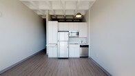 This single-floor unit combines modern design with industrial character, creating a clean and functional living space. The kitchen is a standout feature, outfitted with sleek white cabinetry extending to the ceiling for ample storage, stainless steel appliances including a dishwasher and microwave, and a compact cooktop with sink. A pendant light fixture hangs from the striking exposed coffered ceiling above, which adds architectural depth and an industrial aesthetic throughout the main living area. The flooring transitions between warm wood-tone planks in the kitchen and living spaces and lighter tones near the entry, lending visual variety across the unit. The open living area is bathed in abundant natural light, courtesy of a generous wall of floor-to-ceiling windows with dark frames that frame sweeping panoramic city views — a truly defining element of the space. The crisp, neutral wall tones throughout keep the interior feeling bright and airy. The bathroom is neatly tucked away and accessible via a private hallway, featuring light wood-tone flooring and clean, minimalist finishes. Wall-mounted sconces provide warm accent lighting near the entry corridor. Overall, the space delivers a well-balanced blend of urban industrial style and contemporary minimalism.