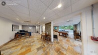 Located at 2517 Guess Rd in Durham, North Carolina, this single-floor amenity space offers a thoughtfully designed environment that blends functionality with a welcoming social atmosphere. The interior is unified by warm-toned natural stone tile flooring throughout, lending the space an earthy, grounded aesthetic that complements its contemporary design.

The main lounge area is anchored by a plush tufted sectional sofa in rich caramel leather, positioned beneath a wall-mounted flat-screen TV and accented by framed artwork and lush indoor greenery. Adjacent to the lounge, a billiards table takes center stage in a game area bathed in natural light from expansive floor-to-ceiling windows that offer open views of the surrounding landscape. A foosball table adds an additional recreational option nearby.

A cozy dining nook features a round wooden table with mid-century-style chairs, nestled beside a stone-clad structural column that adds a rustic architectural detail. The space also includes a dedicated reception area with a sleek dark-wood desk, guest seating, and a private office room — ideal for administrative use or resident services.

The drop ceiling with recessed and panel lighting ensures consistent, even illumination throughout. Neutral gray walls, clean lines, and curated décor create a polished yet relaxed ambiance suited for both leisure and day-to-day interactions.