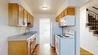 This two-floor residence offers a well-organized layout designed for practical, comfortable living. The galley-style kitchen is equipped with warm honey-toned wood cabinetry lining both walls, a double-basin sink, laminate countertops, and white appliances including an electric range, over-the-range microwave, refrigerator, and dishwasher. Durable hardwood-style flooring runs throughout the kitchen and entry areas, adding warmth to the space. A central staircase with a classic black wood railing and carpeted treads connects the two floors, with the kitchen visible from the base of the stairs. The main living area is generously sized, featuring plush neutral carpeting and a large sliding glass door dressed with vertical blinds that fills the room with natural light and provides a view of the outdoors. Multiple bedrooms are found throughout the home, each finished with soft carpeting and individual closets — some featuring built-in shelving for added storage. The bedrooms are bright and airy, with windows allowing in natural light. The clean, neutral color palette of white walls and beige carpet carries consistently through each room, lending the home a cohesive and move-in-ready appeal. Textured ceilings add a subtle architectural detail throughout the upper and lower levels.