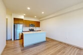 Located at 430 W Stuart Rd in Bellingham, WA, this well-appointed two-bedroom, two-bathroom unit offers a thoughtfully designed single-level layout with a clean, contemporary aesthetic. The open-concept kitchen and living area serves as the heart of the home, anchored by a large central island with a white quartz countertop and an undermount sink. The kitchen is outfitted with warm wood-toned cabinetry, stainless steel appliances including a refrigerator, microwave, and range, and a white tile backsplash that adds a crisp contrast. Light wood-style luxury vinyl plank flooring extends throughout the main living areas, lending warmth and continuity to the space. The living room receives natural light through a wide window, creating a bright and welcoming atmosphere. A functional hallway connects the main living area to the two bedrooms and bathrooms. The primary bedroom features wood-style flooring and a window with outdoor views, while the secondary bedroom is carpeted and also benefits from natural light. Both bedrooms include closet storage. The bathrooms are finished with wood-toned vanities, white quartz countertops, and full tub-shower combinations. Recessed lighting throughout the unit provides consistent, modern illumination.