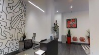 This single-floor commercial space presents a well-appointed reception area defined by its contemporary design aesthetic. The room features notably high ceilings that amplify the sense of openness, with crisp white walls serving as a clean backdrop throughout. A sleek, dark reception desk anchors the space, complemented by a black ergonomic chair and organized workstation equipment, signaling a professional, functional environment.

Illumination is provided by a combination of a suspended linear pendant light and recessed ceiling spotlights, creating a well-balanced and modern lighting scheme. The dark hardwood flooring grounds the space and adds warmth against the predominantly white interior.

Artwork plays a prominent role in defining the character of the space. A bold black-and-white mural-style graphic adorns one wall, while a vivid pop-art floral print featuring red flowers commands attention on the opposite side. Red accent pieces, including a sculptural chair and planter, carry this color theme throughout. Tropical floral arrangements and lush greenery add natural freshness to the environment, completing a polished, gallery-like reception space.