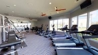This fitness center is a comprehensive, single-level facility designed to support a full range of workout routines. The space is anchored by a row of treadmills and elliptical machines positioned along expansive floor-to-ceiling windows, offering bright natural light and pleasant outdoor views of a pool area and surrounding greenery. Ceiling fans with red-accented blades are mounted throughout, ensuring comfortable air circulation during workouts. The flooring features a distinctive wave-patterned carpet that adds a modern aesthetic to the space. A dedicated free weights area lines one wall, equipped with multiple dumbbell racks offering a wide range of weights, a medicine ball rack, and a full-length mirror that enhances the sense of space and supports proper form. The strength training section includes multiple cable and resistance machines, along with stability balls, catering to a variety of exercise disciplines. Recessed lighting and crown molding along the ceiling contribute a polished, finished look to the interior. Wall-mounted televisions provide entertainment for users during cardio sessions. The overall ambiance is bright, open, and functional, making this fitness center a well-rounded amenity suitable for both casual exercisers and dedicated fitness enthusiasts.