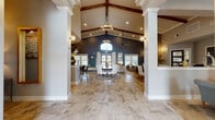 This single-story space makes a striking architectural statement through its dramatic vaulted ceiling, where rich exposed wood beams contrast beautifully against crisp white trim and warm neutral tones. Tall white columns frame the expansive main gathering area, drawing the eye toward a bold dark accent wall anchored by an arched transom window and double glass doors that invite natural light deep into the interior. The main hall is grounded by large-format stone-look tile flooring and centered around a round dining table with upholstered white chairs, set atop a layered area rug. Statement chandeliers and globe wall sconces provide layered ambient lighting throughout. A comfortable lounge area to one side features plush sofas, a glass-top coffee table, and framed wall art beneath the soaring ceiling. A dedicated office area offers dark desks paired with contemporary seating, ideal for business or leasing functions. The kitchen features a long stone-topped bar with dark bar stools, white shaker cabinetry, a patterned tile backsplash, stainless steel appliances, and dramatic pendant lighting, completing this well-appointed and versatile space.