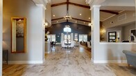 This single-story space makes a striking architectural statement through its dramatic vaulted ceiling, where rich exposed wood beams contrast beautifully against crisp white trim and warm neutral tones. Tall white columns frame the expansive main gathering area, drawing the eye toward a bold dark accent wall anchored by an arched transom window and double glass doors that invite natural light deep into the interior. The main hall is grounded by large-format stone-look tile flooring and centered around a round dining table with upholstered white chairs, set atop a layered area rug. Statement chandeliers and globe wall sconces provide layered ambient lighting throughout. A comfortable lounge area to one side features plush sofas, a glass-top coffee table, and framed wall art beneath the soaring ceiling. A dedicated office area offers dark desks paired with contemporary seating, ideal for business or leasing functions. The kitchen features a long stone-topped bar with dark bar stools, white shaker cabinetry, a patterned tile backsplash, stainless steel appliances, and dramatic pendant lighting, completing this well-appointed and versatile space.