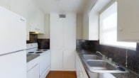 This single-floor space includes a galley-style kitchen designed for efficient use of space. The kitchen features white cabinetry lining both walls, providing ample storage through upper and lower cabinets, as well as a tall pantry-style cabinet unit that extends to the ceiling, offering additional storage capacity. The countertops display a dark speckled finish, complemented by matching dark tile backsplashes that create a cohesive, classic aesthetic throughout the cooking area. A white electric range with coil burners sits along one wall beneath a range hood, while a double-basin stainless steel sink is positioned on the opposite counter, conveniently located near a window that allows natural light to filter into the space. The flooring consists of terracotta-toned tiles, adding warmth to the kitchen's overall appearance. A ceiling vent ensures proper ventilation throughout the area. The layout and storage solutions make this kitchen practical and straightforward, suited for everyday cooking needs.