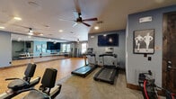 Located in Rockwall, TX, this fitness center offers a thoughtfully designed single-floor exercise space that accommodates a wide range of workout styles. The interior is finished with warm hardwood flooring throughout the open areas, complemented by light blue and gray walls that create a clean, energizing atmosphere. Recessed lighting and ceiling fans ensure the space remains well-lit and comfortable during use.

The cardio zone is equipped with multiple treadmills and stationary cycling machines, each featuring individual consoles, and a wall-mounted flat-screen television for entertainment during workouts. A dedicated open studio area along one wall features a full-length mirrored surface paired with mounted ballet barres, making it well-suited for barre classes, stretching, and flexibility training. A dumbbell rack with a range of weights is positioned nearby for strength training convenience.

Rustic-style wood-trimmed doorframes add a distinctive architectural character to the space, providing a warm contrast to the modern fitness equipment. Natural light filters in through windows, adding brightness to the environment. The layout is organized to provide ample open floor space for group exercise or individual training sessions. A seating area with chairs offers a rest zone, and sanitation stations are conveniently placed throughout. This fitness center is a comprehensive facility designed to support diverse fitness goals.