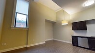 Unit 303 is a well-appointed 1-bedroom, 1-bathroom residence situated on the third floor. The living area immediately draws attention with its notably elevated ceilings, which lend an airy, open quality to the space. Warm, neutral green-toned walls provide a versatile backdrop that complements a variety of décor styles. The flooring throughout features durable hardwood-style planks in a rich, dark tone, adding a sense of warmth and continuity across the space. A large window along one wall allows natural light to filter in, illuminating the room and offering views of the surrounding exterior. A centrally hung drum pendant light fixture provides functional and stylish overhead illumination. The layout flows naturally from the living area toward the kitchen, which is partially visible and features dark cabinetry, hinting at a cohesive, modern design aesthetic. Architectural details such as clean white crown molding and baseboards provide crisp contrast against the muted wall tones. The overall ambiance of the unit is calm and functional, offering a comfortable single-floor living experience with thoughtful design elements throughout.