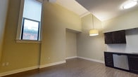 Unit 303 is a well-appointed 1-bedroom, 1-bathroom residence situated on the third floor. The living area immediately draws attention with its notably elevated ceilings, which lend an airy, open quality to the space. Warm, neutral green-toned walls provide a versatile backdrop that complements a variety of décor styles. The flooring throughout features durable hardwood-style planks in a rich, dark tone, adding a sense of warmth and continuity across the space. A large window along one wall allows natural light to filter in, illuminating the room and offering views of the surrounding exterior. A centrally hung drum pendant light fixture provides functional and stylish overhead illumination. The layout flows naturally from the living area toward the kitchen, which is partially visible and features dark cabinetry, hinting at a cohesive, modern design aesthetic. Architectural details such as clean white crown molding and baseboards provide crisp contrast against the muted wall tones. The overall ambiance of the unit is calm and functional, offering a comfortable single-floor living experience with thoughtful design elements throughout.