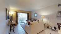 This single-floor residence offers a thoughtfully designed layout that balances comfort and functionality across its key living areas. The living room serves as the heart of the home, featuring light-toned tile flooring, neutral walls with warm yellow accents, and a large sliding glass door fitted with vertical blinds and plaid draping curtains that open to an outdoor patio. Natural light fills the space, complementing the plush sectional seating, a mosaic-top coffee table, and a decorative circular mirror that adds a refined touch to the wall.

Adjacent to the living room, the dining area provides an intimate setting centered around a round bistro-style table with wrought iron chairs, illuminated by a pendant light overhead. The kitchen is well-equipped, featuring crisp white cabinetry, granite-style countertops, a stainless steel sink, and black appliances including a dishwasher and refrigerator, all set over easy-clean tile flooring.

The bedrooms are carpeted for warmth and comfort, each offering generous proportions, natural window light with decorative curtains, and ample closet space — one room notably features a walk-in closet. The bathroom presents a granite-topped vanity, a large wall mirror with overhead lighting, and tile flooring, completing this well-rounded residence.