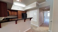 This two-floor residence offers a well-organized and functional layout designed with everyday comfort in mind. The kitchen is a standout feature, showcasing rich warm-toned wood cabinetry paired with dark granite-style countertops that create a classic contrast. A breakfast bar with decorative corbel supports extends from the counter, providing a casual dining or workspace area. Black appliances, including a full-size refrigerator, add a sleek, cohesive look to the kitchen. Crown molding runs along the ceiling throughout, lending an elevated, polished character to the interiors. Adjacent to the kitchen, a hallway leads further into the home, flanked by louvered bifold doors that likely conceal a utility or storage closet — a practical touch that keeps the space tidy. The flooring transitions from hard-surface wood-look planks in the kitchen and hallway areas to plush carpet in the living spaces, offering both durability and warmth where it matters most. Neutral beige and warm white tones on the walls and trim create a bright, inviting atmosphere that flows naturally throughout both levels of the home.