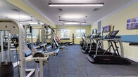 Located at 89 Privilege Rd in Bloomfield, CT, this fitness center is a comprehensively outfitted single-floor facility designed to support a wide range of workout routines. The space features light yellow walls accented by a blue horizontal band, giving the room a clean, energetic atmosphere. Multi-panel fluorescent lighting runs across the ceiling, ensuring consistent, bright illumination throughout.

The cardio section is anchored by a row of LandIce treadmills and elliptical machines, each equipped with individual digital consoles. Wall-mounted flat-screen televisions are positioned along the cardio zone for entertainment during workouts. On the opposite side of the floor, a multi-station cable and weight machine provides options for comprehensive strength training, complemented by an adjustable bench.

A dedicated free-weight area includes a full dumbbell rack with varying weights, an adjustable workout bench, stability balls, and a tiered medicine ball rack ranging in multiple weights. Windows along the far wall allow natural light to filter in, offering views of the surrounding outdoor greenery. The floor is covered in durable, multi-toned carpet tiles suited for high-traffic use. A coat rack near the entry door adds a practical touch. Overall, the space is organized, functional, and well-maintained.