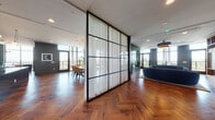 This expansive single-floor space is defined by a cohesive design language that blends contemporary elegance with warm, organic materials. Rich herringbone-patterned hardwood flooring flows throughout, creating a visual thread that unifies each area. The entry is marked by striking teal-painted double glass doors with paneled millwork, setting a refined tone from the moment of arrival.

The open layout connects several distinct living zones. A dramatic floor-to-ceiling glass panel room divider with sheer white curtains and a black steel grid frame delineates spaces while preserving sightlines and natural light. The living area features expansive floor-to-ceiling windows that frame sweeping outdoor views, complemented by a curved sofa, statement pendant lighting, and a built-in wood bookcase. A second seating area mirrors this openness with additional glazed doors leading to an outdoor terrace.

The kitchen and dining zone showcases deep green cabinetry, a sleek dark granite island, brass pendant fixtures, and a large dining table surrounded by warm-toned upholstered chairs. Throughout, recessed lighting, bold accent walls in forest green and charcoal, and curated furnishings contribute to a polished, upscale ambiance.