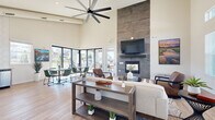 Located at 300 Canvas Ridge Ave in Castle Rock, Colorado, this single-story community clubhouse offers a thoughtfully designed gathering space characterized by soaring ceilings and an abundance of natural light. The main lounge area serves as the heart of the space, anchored by a dramatic floor-to-ceiling stone tile fireplace with a built-in gas hearth and wall-mounted television above. Oversized ceiling fans complement the open volume of the room, while large-format sliding glass doors and clerestory windows flood the interior with daylight and provide views of the surrounding outdoor patio and community. Light wood-look flooring flows throughout, tying together the open-concept layout. Comfortable seating areas with neutral sofas, leather accent chairs, and contemporary coffee tables create inviting spots for lounging, while a separate dining zone with modern tables and green upholstered chairs accommodates casual gatherings. The kitchenette is well-appointed with stainless steel appliances including a side-by-side refrigerator and dishwasher, granite countertops, gray cabinetry, open shelving, and a striking hexagonal white tile backsplash. Together, these spaces deliver a warm, modern ambiance suited for both relaxed socializing and community events.