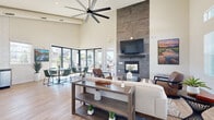 Located at 300 Canvas Ridge Ave in Castle Rock, Colorado, this single-story community clubhouse offers a thoughtfully designed gathering space characterized by soaring ceilings and an abundance of natural light. The main lounge area serves as the heart of the space, anchored by a dramatic floor-to-ceiling stone tile fireplace with a built-in gas hearth and wall-mounted television above. Oversized ceiling fans complement the open volume of the room, while large-format sliding glass doors and clerestory windows flood the interior with daylight and provide views of the surrounding outdoor patio and community. Light wood-look flooring flows throughout, tying together the open-concept layout. Comfortable seating areas with neutral sofas, leather accent chairs, and contemporary coffee tables create inviting spots for lounging, while a separate dining zone with modern tables and green upholstered chairs accommodates casual gatherings. The kitchenette is well-appointed with stainless steel appliances including a side-by-side refrigerator and dishwasher, granite countertops, gray cabinetry, open shelving, and a striking hexagonal white tile backsplash. Together, these spaces deliver a warm, modern ambiance suited for both relaxed socializing and community events.