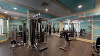 This fitness center offers a comprehensive workout environment spread across a single floor, designed with both functionality and aesthetic appeal in mind. The space is finished in a distinctive teal and cream color scheme, with tray ceilings featuring recessed lighting that illuminate the rubber-floored workout areas throughout. Large wall-to-wall mirrors line key sections of the gym, visually expanding the space and allowing users to monitor their form during exercise.

The main workout area is anchored by a multi-station Magnum cable machine and is complemented by a full dumbbell rack with adjustable benches, accommodating a wide range of free weight exercises. A dedicated strength zone houses plate-loaded equipment, including a leg press and additional Magnum machines, catering to lower and upper body training. A secondary room features built-in storage cubbies stocked with kettlebells and exercise mats, alongside a vertical knee raise station for core work.

Natural light filters into one section through slatted blinds, adding brightness to the space. The entry area features wall-mounted informational displays and a security camera, reflecting a well-managed facility. Overall, the gym presents a clean, organized, and fully equipped environment suitable for a broad range of fitness routines.