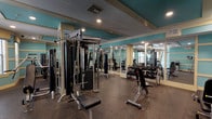 This fitness center offers a comprehensive workout environment spread across a single floor, designed with both functionality and aesthetic appeal in mind. The space is finished in a distinctive teal and cream color scheme, with tray ceilings featuring recessed lighting that illuminate the rubber-floored workout areas throughout. Large wall-to-wall mirrors line key sections of the gym, visually expanding the space and allowing users to monitor their form during exercise.

The main workout area is anchored by a multi-station Magnum cable machine and is complemented by a full dumbbell rack with adjustable benches, accommodating a wide range of free weight exercises. A dedicated strength zone houses plate-loaded equipment, including a leg press and additional Magnum machines, catering to lower and upper body training. A secondary room features built-in storage cubbies stocked with kettlebells and exercise mats, alongside a vertical knee raise station for core work.

Natural light filters into one section through slatted blinds, adding brightness to the space. The entry area features wall-mounted informational displays and a security camera, reflecting a well-managed facility. Overall, the gym presents a clean, organized, and fully equipped environment suitable for a broad range of fitness routines.